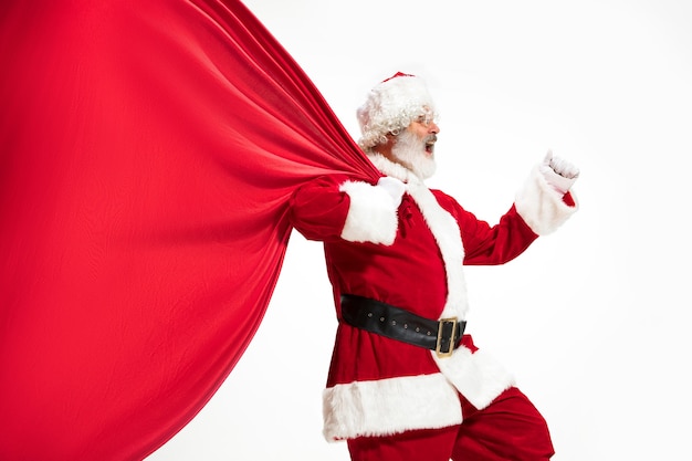 산타 클로스는 흰색 배경에 고립 된 크리스마스 선물로 가득 찬 거대한 가방을 당기고 있습니다. 전통 의상을 입은 백인 남성 모델. 2020년 새해, 선물, 휴일, 겨울 분위기. 귀하의 광고에 대 한 Copyspace입니다.