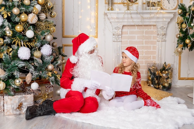 Santa Claus reads a book to a child girl at the Christmas tree