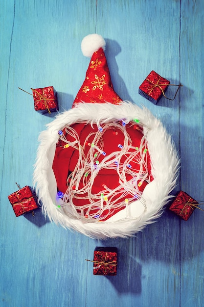 Santa Claus's hat has a lighted garland and presents around it, on a blue wooden background.