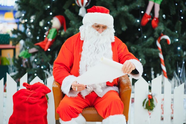 Santa Claus sitting on the background of the Christmas tree in the lobby of the modern shopping mall. Bokeh basic background for design.