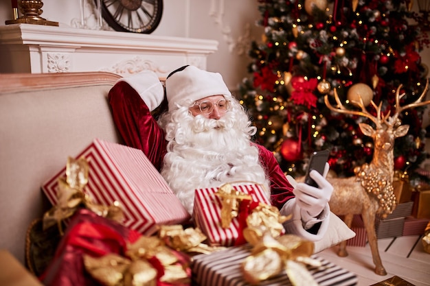 Santa Claus sitting on couch and talking on mobile phone near the fireplace and christmas tree with gifts New year and Merry Christmas happy holidays concept