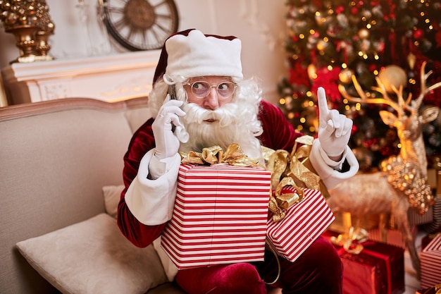 Santa Claus sitting on couch and talking on mobile phone near the fireplace and christmas tree with gifts. New year and Merry Christmas , happy holidays concept