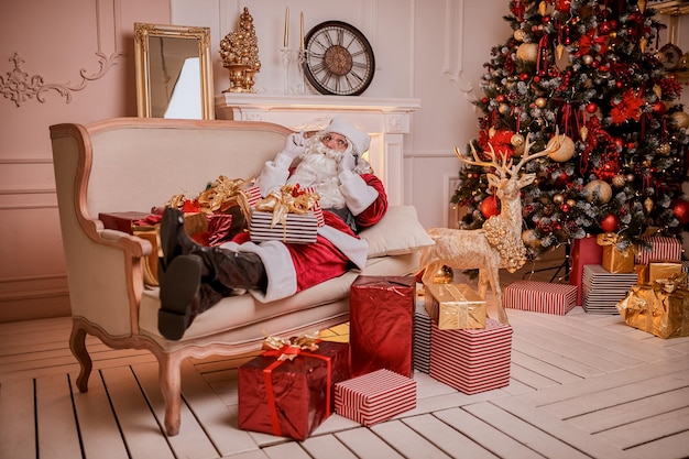 Санта-Клаус сидит на диване и разговаривает по мобильному телефону возле камина и елки с подарками. Новый год и счастливого Рождества, концепция счастливых праздников