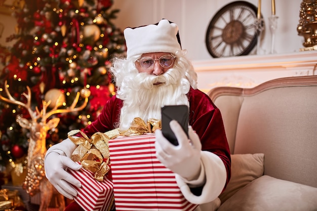Санта-Клаус сидит на диване и разговаривает по мобильному телефону возле камина и елки с подарками.