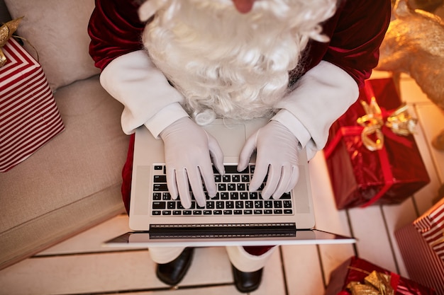 Santa Claus sitting at his home and reading email on laptop with Ñhristmas requesting or wish list near the fireplace and tree with gifts. New year and Merry Christmas , happy holidays concept