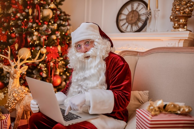 Babbo natale seduto a casa sua e leggendo e-mail sul laptop con ñ richiesta di natale o lista dei desideri vicino al camino e albero con regali. anno nuovo e buon natale, concetto di buone feste