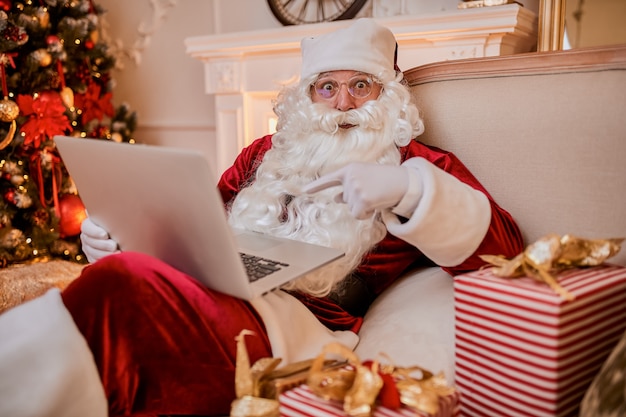 Санта-Клаус сидит у себя дома и читает электронную почту на ноутбуке с запросом на рождество или списком желаний возле камина и елки с подарками. Новый год и счастливого Рождества, концепция счастливых праздников