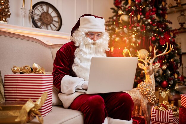 Santa Claus sitting at his home and reading email on laptop with shristmas requesting or wish list near the fireplace and tree with gifts New year and Merry Christmas happy holidays concept