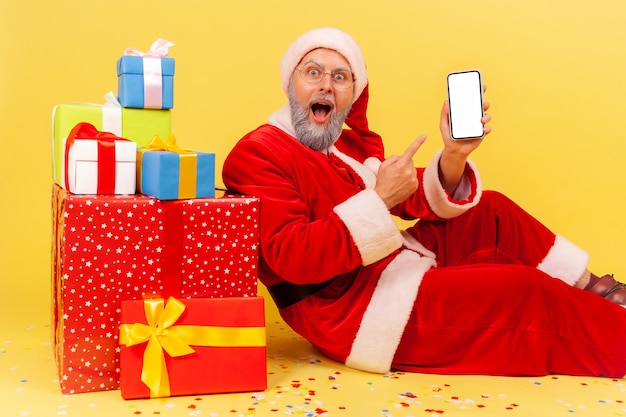 Santa claus sitting pointing white blank screen of smart phone, surrounded with Christmas gifts.