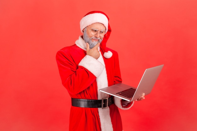 Santa claus staande met kin, kijkend naar laptopscherm met peinzende uitdrukking.