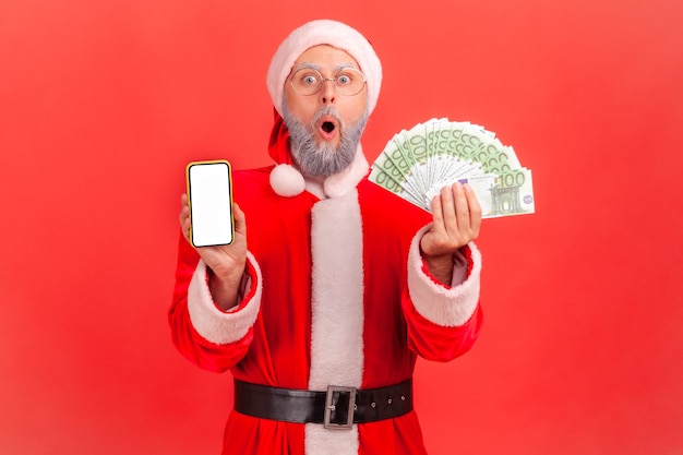 Santa claus standing with open mouth, showing euro banknotes and cell phone with empty display.