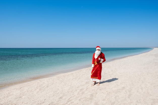 Foto babbo natale con un sacco di regali che cammina sulla spiaggia sabbiosa - immagine