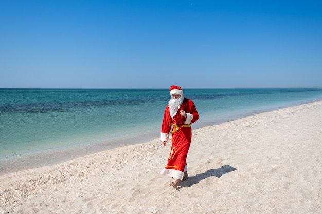 Photo santa claus with a bag of gifts walking on the sandy beach. turquoise water. copy space for your text - image