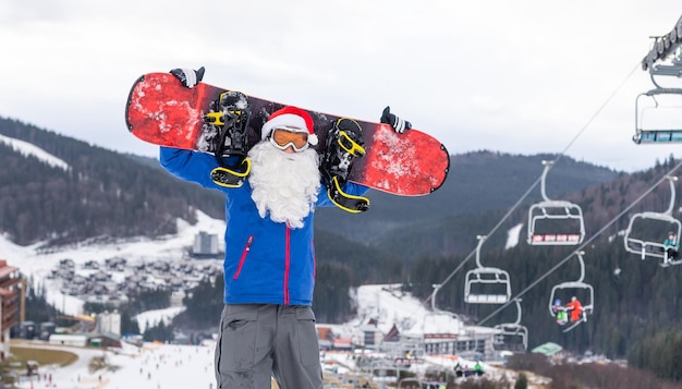 雪に覆われた冬の山のスキーリゾートの風景と青い空、新年またはクリスマスのグリーティングカードに対してスノーボードとサンタクロース。