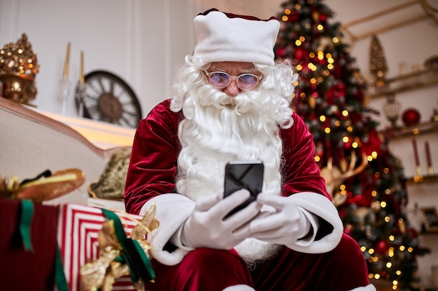 Santa claus zittend op de bank en praten op mobiele telefoon bij de open haard en kerstboom met geschenken. nieuwjaar en vrolijk kerstfeest, gelukkig vakantieconcept