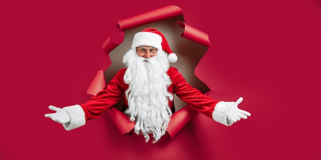 Санта-Клаус заглядывает в отверстие на красной бумаге, жестикулируя руками, как приветственная новогодняя реклама
