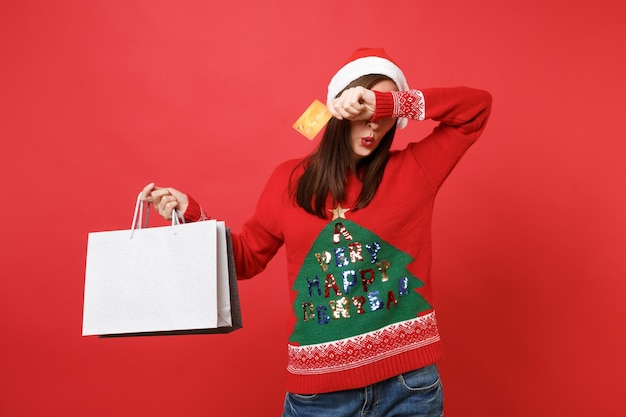 Santa girl covering eyes with hand hold credit card, packages bags with purchases after shopping isolated on red background. Happy New Year 2019 celebration holiday party concept. Mock up copy space.