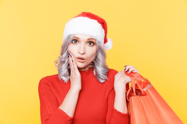 Santa girl in hat holding big bags with presents isolated in yellow room.