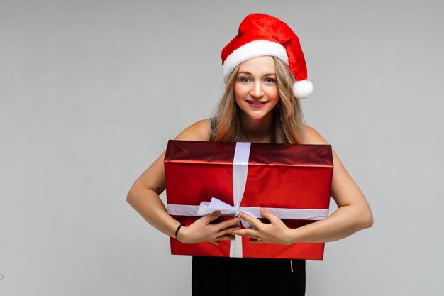 Santa girl in red hat with large festive gift smiling posing on gray background with copy space for xmas new year ad