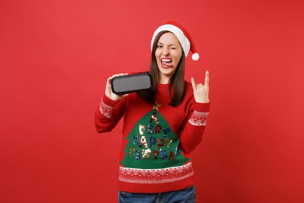 Santa girl showing horns gesture, depicting heavy metal rock sign, holding portable wireless bluetooth music speaker isolated on red background. Happy New Year 2019 celebration holiday party concept.