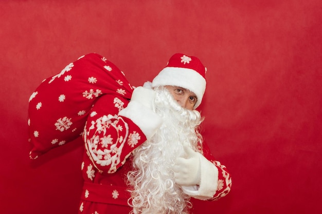 Santa holds a bag of gifts on a red background behind his back and gives a thumbs up