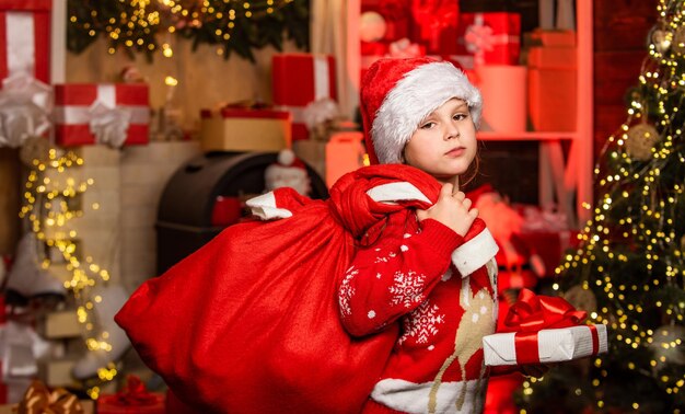 Santa kid with presents Little santa girl at christmas tree What is in her bag Decorate home with joy Xmas mood Holiday celebration Happy new year Child in santa hat carry heavy bag on back