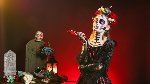 Santa muerte model talking on smartphone call, wearing\
halloween make up and flowers crown in studio. la cavalera catrina\
using smartphone to chat on dios de los muertos mexican\
holiday.