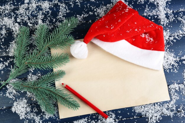 Santa red hat with branch of fir-tree, sheet of paper, pencil and snowflakes on color wooden table