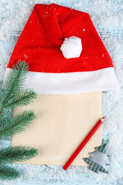 Santa red hat with fir-tree branch, sheet of paper, pencil  and snowflakes on color wooden surface