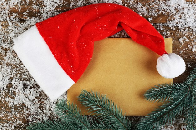 Santa red hat with snowflakes, sheet of paper and branch of fir-tree on wooden surface