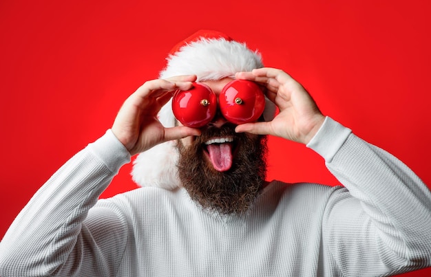 Santa shows tongue. Happy Santa hold christmas balls. Bearded man holds Christmas toy near eyes. Christmas mood. Happy holidays. Santa's hat. Funny faces.