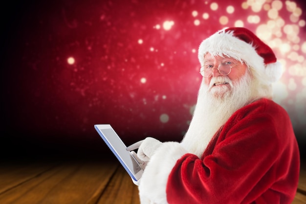 Santa uses a tablet PC and smiles into the camera against shimmering light design over boards