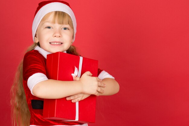 Foto santaâ € ™ s kleine helper. studio portret van een klein schattig meisje draagt kerst outfit met een geschenk en vrolijk lachen op rode copyspace aan de zijkant