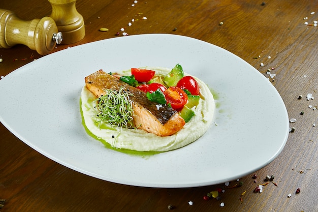 Sappige zalm steak op een witte plaat met een bijgerecht van aardappelpuree, avocado en cherrytomaatjes met groene saus. Houten tafel. Sluit omhoog op smakelijke restaurantzeevruchten