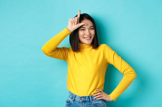 Sassy asian girl mocking lost team, showing loser sign on forehead and smiling pleased