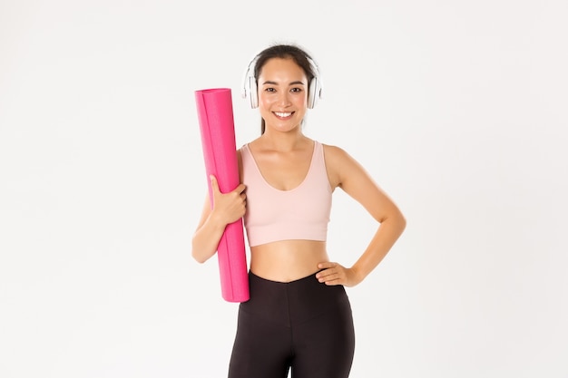 Sassy bella ragazza asiatica fitness, donna pronta per lezioni di yoga o allenamento a casa, con tappetino in gomma per allenamento e ascolto di musica in cuffia.