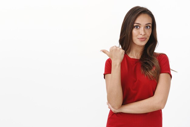 Sassy gorgeous brunette female in red tshirt having chat with friends discuss handsome guy standing behind pointing thumb left and pouting impressed smiling pleased near white background