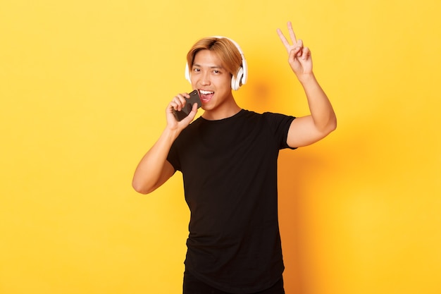 Sassy handsome asian guy in headphones, playing karaoke app, singing into mobile phone microphone, showing peace gesture, standing yellow wall