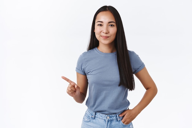 Sassy knappe zelfverzekerde brunette aziatische vrouw die advies geeft welke productkeuze, beste keuze toont, naar links wijst, tevreden grijns geeft wetende blik, hand in jeanszak houdt, witte muur