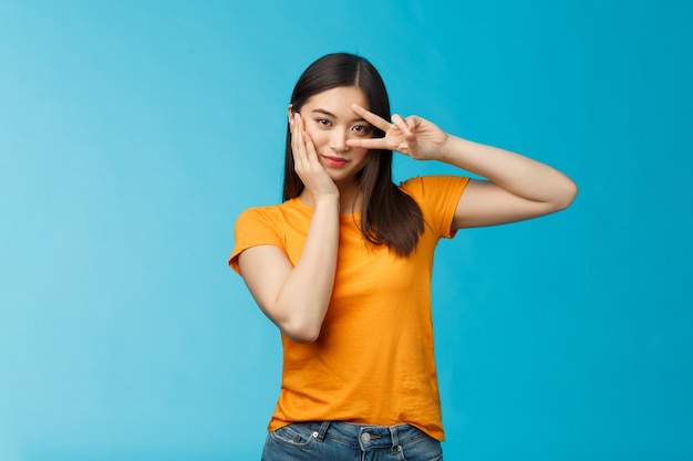 Sassy, stijlvol, goed uitziend Aziatisch assertief Koreaans meisje toont vredesoverwinningsteken op oog, raak wang aan, kijk krachtig en gemotiveerd, staande brutale kokette blauwe achtergrond, draag geel t-shirt