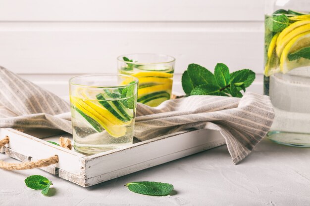 Sassy water for detox infused with lemon, cucumber and mint in glasses at white wooden tray. Healthy lifestyle concept.