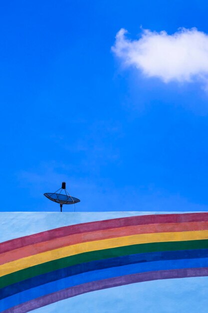 Satellite dish on rooftop with rainbow stripes on building wall surface against blue sky background