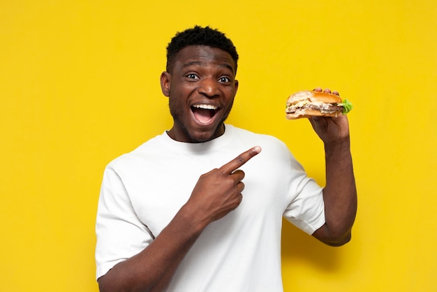 Satisfied african american man in white tshirt holding big burger and pointing with his hand