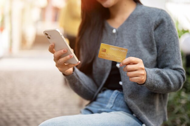 a satisfied asian woman sitting on a bench outdoors holding her smartphone and a credit card