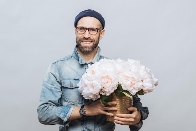 Satisfied bearded male with cheerful expression has broad smile holds bunch of flowers wears hat and denim jacket isolated over white background Satisfied smiling man poses indoor with flowers