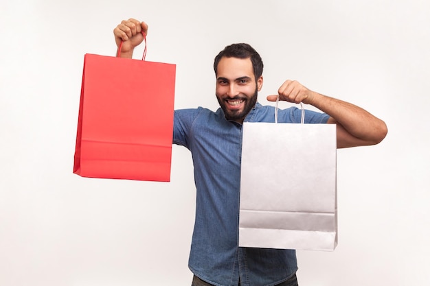 Satisfied bearded man in blue shirt bragging with paper shopping bags, satisfied with black friday big sale. Indoor studio shot isolated on white background