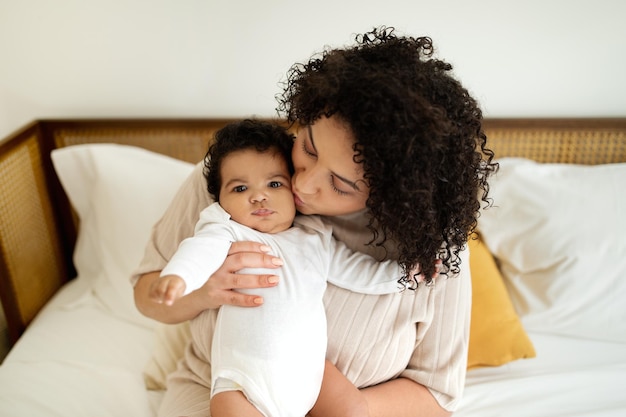 Satisfied black millennial mom kiss little baby sit on white bed enjoy love comfort and rest