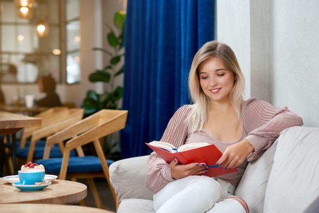 Satisfied blonde girl reading interesting book sitting in cafe.