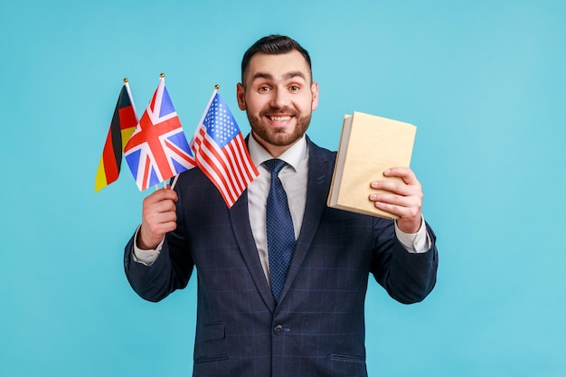 Satisfied businessman or teacher wearing official style suit, holding flags of Germany, Great Britain and USA and book, looking at camera with smile. Indoor studio shot isolated on blue background.