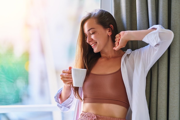 Satisfied carefree casual young beautiful happy smiling woman\
drinking coffee in lazy morning at comfy home interior